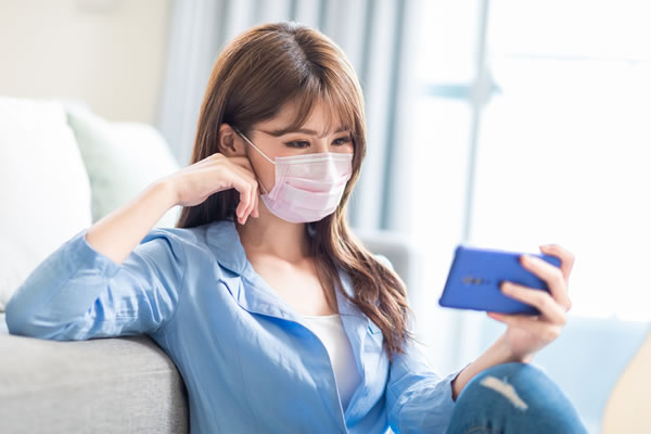 woman wearing mask sitting on couch reading cell phone